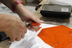A decorative photo showing hands cutting orange class for a Stained Glass project.