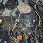 A detailed, high-angle close-up shot of a denim textile artwork, showcasing a dense, layered composition of stylized flowers and botanical motifs. The background is a rich patchwork of various shades of blue denim, with visible seams and scattered white "sashiko-style" stitching, creating texture. Prominent features include: - Two large, stylized poppy-like forms in the upper centre: one dark blue with white outlining, and one lighter, faded blue with subtle stitching, both on long, narrow stems. - Numerous smaller embroidered and appliquéd flowers in various shades of blue, black, and white, some with orange or yellow centres. - Ribbons of fabric, resembling stems or vines, in shades of grey and black, intertwine through the design. Some of these strips appear to be zippers, with visible teeth and pulls integrated into the art. - Delicate white embroidered leaf and floral patterns are visible throughout the darker denim sections. The image highlights the intricate craftsmanship and the creative repurposing of denim and other materials in the textile art piece.