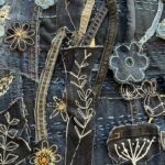 A detailed, high-angle close-up shot of a denim textile artwork, showcasing a rich array of embroidered and appliquéd floral and plant motifs. The background is a patchwork of various shades of dark blue denim, some with visible seams and light blue or white stitching that adds texture. The artwork features numerous stylized elements: - Several dark blue floral appliques are outlined in white stitching, some with yellow or orange centers. - White embroidered outlines create delicate leaf and stem patterns, as well as abstract floral shapes. - Strips of denim, some frayed or with visible yellow stitching, are used as dimensional elements, possibly representing stems or vines, curving through the composition. - Small, light blue denim floral appliques are scattered throughout. The intricate stitching and layered denim pieces create a detailed and textured surface, emphasizing the handcrafted nature of the art.