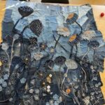 A high-angle, full shot captures a large, rectangular textile artwork laid flat on a light wooden table. The artwork is crafted from various shades of blue denim, pieced together to form a rich, textured background. The surface is extensively embellished with appliqué and embroidery, depicting a vibrant and detailed field of stylized flowers and seed pods. Key elements include: - Several large, dark blue seed pods, some with textured surfaces, and lighter greyish-white poppy-like flowers with orange or black centres. - Numerous smaller flowers in different shades of blue and black, outlined with white stitching, and accented with orange or yellow. - Thin, dark stems made from denim strips or embroidery connect many of the floral elements. - The lower section features a dense array of intricate white embroidered patterns, resembling small flowers, leaves, and abstract botanical forms. - A few names, such as "Jenny", are visible in white stitching on the lower left portion of the artwork. Two black chairs are partially visible behind the table at the top of the image. A small red ball is also visible on the table to the right. The image showcases the entirety of the elaborate denim art piece.