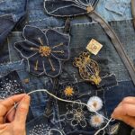 A high-angle close-up shot shows two hands working on a denim textile art piece. The background is a rich patchwork of various shades of blue denim, with visible seams and different textures. The artwork is embellished with intricate embroidery and appliqué. Prominent elements include: - A large, dark blue floral appliqué with white outline stitching and orange details in the upper centre. - A dark blue seed pod appliqué with gold stitching. Several smaller embroidered flowers with white petals and orange or white centres. - White stitching forms starburst patterns and other decorative motifs across the denim. - A grey zipper is integrated into the denim patchwork on the right side. - A small, light brown tag with "33" in black is visible near the top right of the seed pod. The hands are holding white yarn or thick thread, actively engaged in the process of adding to the textile, with loops of the thread resting on the artwork. The image highlights the detailed craftsmanship and the hands-on creation of the art piece.