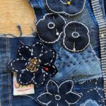 A detailed, close-up, and slightly overhead shot of a dark blue denim textile, showcasing a section of an embroidered and appliquéd artwork. The denim has visible seams and varying shades of blue, indicating it is made from repurposed fabric. Several dark blue floral appliques are prominent, each outlined with white stitching. Some of these flowers are pinned to the background denim with white or yellow-headed pins, suggesting they are in the process of being attached. One large flower in the centre features intricate white and orange stitching for its petals and centre. The background denim also has white stitched patterns, including dashed lines and small decorative motifs. A striped fabric piece, possibly a zipper or trim, runs vertically along the right side of the image. A small, light-coloured fabric tag is partially visible under one of the flowers on the left. The image emphasizes the textured and handcrafted nature of the textile art.