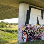 A large concrete support column of a bridge is covered in vibrant graffiti, standing next to a grassy, open field under the bridge. The column's upper section has large black, angular shapes, while its base is covered in colourful, intricate graffiti tags and characters. To the left, the underside of the bridge stretches across the top of the frame. Beyond the column, a bright green field with scattered trees extends into the distance, and the horizon is visible with a clear sky.