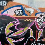 A dynamic close-up of a graffiti-covered wall, filled with vibrant colours and abstract shapes. In the foreground, bold black forms are outlined with neon pink and lime green, creating a chaotic, energetic pattern. Above this, the background features lighter, more varied elements: a white dice with black dots, a white abstract shape resembling a flame or horn, and various other shapes and colours including orange, light blue, pink, and hints of yellow and white circles. A stylized face with white outlines and a dark eye socket is visible on the upper right, and another dark, somewhat blocky face is in the upper left corner. The overall impression is one of complex and layered street art.
