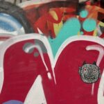 A close-up shot of a concrete wall covered in colourful graffiti. In the foreground, there are two large, rounded shapes outlined in white and filled with a deep reddish-pink. On the rightmost red shape, a black and white sticker or stencil of a stylized monster face with small horns, large eyes, and a jagged mouth is prominently displayed. In the background, other spray-painted elements are visible, including bright turquoise blobs, red and orange abstract shapes, and hints of blue and white.