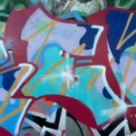 A vibrant close-up of a graffiti-covered wall, showcasing an overlapping array of spray-painted shapes and colours. Dominant elements include bold, dark red outlines, light blue and teal fills, and intricate yellow and white highlights that appear to form letters or abstract designs. A darker blue shape is visible on the upper right, contrasting with lighter pink and orange hues in the background. The texture of the wall is also visible beneath the layers of paint.