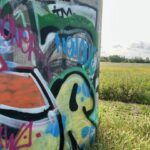 A large, cylindrical concrete pillar, heavily covered in colourful graffiti, stands prominently on the left side of the frame, contrasting with a bright green field that stretches into the distance on the right. The graffiti features various styles and colours, including a large yellow section with blue splatters, a green outline, and bold black shapes. Above it, there are layers of green, black, and blue spray paint, with some visible text in pink and light blue. The field is lush with tall grasses and other vegetation, and the sky above is bright with some clouds visible.
