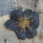 A close-up, high-angle shot focuses on a dark blue denim flower appliqué resting on a lighter denim background. The flower has five rounded petals, cut from dark blue denim, with slightly frayed edges. The centre of the flower is intricately embroidered with a burst of white and orange-gold stitches, resembling stamens. The background is a patchwork of various shades of blue denim, joined with visible seams and adorned with delicate white "sashiko-style" dashed stitching, adding texture and a handcrafted feel. The image highlights the detailed craftsmanship of the textile art.