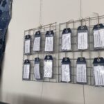 A slightly angled, eye-level shot captures a display of twelve white rectangular tags with information about fashion and its environmental impact. Each tag is suspended by a loop of brown twine from a metal grid. The grid, made of intersecting horizontal and vertical metal bars, is mounted against a plain white wall. To the left, part of a richly embroidered blue textile artwork depicting a tree with birds and floral motifs is visible. The tags are arranged in three rows of four. The tags contain text such as: "The production of one pair of jeans requires approximately 3,000 litres of water." "500,000 tons of microfibres are released into our oceans from washing clothes." "Fashion production doubled between 2000 and 2014." "Around 20% of industrial water pollution worldwide originates from the fashion industry." "The industry uses up 93 billion cubic metres of water annually, enough to meet the consumption needs of five million people." "Fashion and its supply chain are the third largest polluting industry in the world."