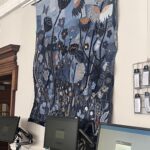An eye-level, slightly angled shot shows a large, vertically oriented textile artwork made from various shades of blue denim, hanging on a plain white wall. The artwork depicts a field of stylized flowers and seed pods in dark blue and black, with accents of orange and white stitching. A butterfly is visible in the upper left. Below the artwork, on a dark surface, are three computer monitors displaying the 'Explore' library login screen. To the far right, partially visible, is a metal grid display of white tags about fashion and its environmental impact.