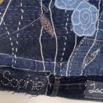 A close-up shot focuses on the lower section of a dark blue denim textile artwork. The texture of the denim is visible, with various shades and seams. The artwork features intricate embroidery and applique. Prominent elements include: - Two light blue, spiral-shaped floral appliques in the upper right. - White dashed lines of stitching that appear to represent stems or grass. - Smaller, detailed orange and brown embroidered leaf-like shapes. - Scattered white and purple stitching creating texture. At the bottom left, a frayed edge of the denim has white embroidery spelling out "Sophie". To the right, another denim tag, also with white embroidery, partially spells out "Isabel". The image highlights the detailed craftsmanship and recycled nature of the materials used in the artwork.