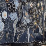 A detailed shot focuses on a vibrant section of a textile artwork crafted from various shades of blue denim. The surface is rich with appliqué and embroidery, forming a dense floral design. Different flowers and leaves are created using varying denim colours, intricate white stitching, and outlines. Some flowers feature orange and gold stitching, while others have black outlines or tiny bead-like accents. A zipper detail is visible at the top, integrating into the design. Along the bottom edge of the artwork, stitched onto denim fragments, are names in white script: "Jacqueline" on the far left, then "Jen", and "Jayne" on the right. The image showcases the textural complexity and collaborative nature of the denim art piece.