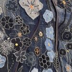 A vibrant, close-up, shot of a denim textile artwork, showcasing an intricate collage of appliqué and embroidery that forms a dense field of flowers. Various shades of blue denim, from light to dark, are pieced together, creating a rich textural background. The flowers are diverse in design: some are outlined in black with white or orange centres, others are made from lighter blue denim with delicate white stitching. Some flowers feature prominent orange or gold stitching for their petals or centres. White embroidered lines, reminiscent of stems or grasses, weave through the floral motifs. Two zips are integrated into the design, running vertically and diagonally through the composition. The stitching and varying textures of the denim are highly detailed, emphasizing the handcrafted nature of the piece.