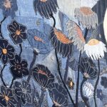 A vibrant, close-up, and slightly overhead shot of a denim textile artwork, showcasing a dense and layered composition of stylized flowers. The background is a patchwork of various shades of blue denim, creating a textured base. The artwork features a variety of floral forms: - Several dark blue flowers with prominent white outlines and orange or gold stitched centres. - Some flowers have a greyish-white or light blue denim base with intricate white and black stitching, often with orange accents, suggesting poppy-like forms. Some of these have a textured, glittery orange or gold fill. - Thin, dark stems extend from many of the flowers. - Smaller, more abstract floral motifs are scattered throughout the composition. - The stitching is highly detailed, adding texture and depth to the recycled denim fabric. The overall impression is a rich, artistic representation of a garden or field of flowers.