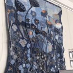 An eye-level, slightly angled shot captures a large, vertical textile artwork made from various shades of blue denim, hanging against a plain white wall. The artwork is richly detailed with appliqué and embroidery, depicting a vibrant field of stylized flowers and seed pods. In the upper section, two prominent dark blue seed pods with textured surfaces are visible, extending on long, curving stems. Below and around them, a multitude of flowers in different shades of blue, black, white, and orange are depicted with intricate stitching. Some flowers have black outlines and orange centres, while others are lighter blue with delicate white embroidery. A butterfly is visible in the upper left. The background is a patchwork of denim, creating a varied texture. Along the bottom edge of the artwork, white stitched names like "Jacqueline" and "Jayne" are visible. In the foreground, the top of two computer monitors can be seen, and to the right, a display grid with white information tags is partially visible.