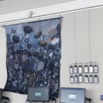 An eye-level, slightly angled shot shows a large, vertically oriented textile artwork made from various shades of blue denim, hanging on a plain white wall. The artwork depicts a field of stylized flowers and seed pods in dark blue and black, with accents of orange and white stitching. A butterfly is visible in the upper left. Below the artwork, on a dark surface, are two computer monitors, both displaying the Explore library login screen. To the right of the artwork, a metal grid display of white tags with information about fashion and its environmental impact is visible. In the foreground, the top of a person's head with blonde hair is partially visible.