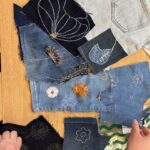 A high-angle shot captures a collection of various denim scraps and embroidered patches laid out on a light wooden surface. The denim pieces range in shade from light blue to dark indigo. Several pieces feature intricate embroidery: - A large dark blue denim patch in the upper left has a white outlined floral or seed pod design with small gold details. - A lighter blue denim patch has a white embroidered leaf design. - Another light blue denim piece displays two round embroidered flowers: one with a white centre and white petals, and another with an orange-gold centre and orange-gold petals. - A dark blue denim patch in the lower left has scattered white embroidered dandelion-like motifs. - Two dark blue denim patches show white outlined floral shapes. - A black and white feathered embroidery is visible on a mid-blue denim piece. - A small dried botanical element is placed on a mid-blue denim piece. I n the lower left, a person's left hand is visible, and in the lower right, part of a person's right hand is visible, holding a striped green and white fabric piece. The image provides a close-up view of the components and work-in-progress of a textile art project.