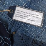 A close-up, high-angle shot focuses on a white rectangular tag with black text, suspended by a loop of brown twine from a metal eyelet. The tag is displayed against a background of patchwork denim fabric, which consists of various shades of blue and features scattered white and light blue "sashiko-style" stitching. The text on the tag reads: "Every second, the equivalent of a rubbish truck load of clothes is burnt or buried in landfill. (Ellen MacArthur Foundation)". Below the tag, a dark blue denim section of the artwork is visible, with light blue embroidered lines forming abstract patterns and cut-out holes creating a textured effect. The image highlights the message about textile waste within the context of a recycled textile art piece.