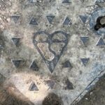 A weathered, square metal utility cover is shown from a high angle, embedded in a gravelly or dirt surface. The cover is adorned with a pattern of raised, triangular studs. In the centre, a distinct embossed symbol of a heart with an abstract, winding shape within it is visible. The metal is discoloured and grimy, indicating age and exposure to the elements. A portion of a dark, braided rope or hose is visible at the bottom left.