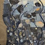 A high-angle, full shot captures a large, rectangular textile artwork laid flat on a grey carpeted surface. The artwork is crafted from various shades of blue denim, pieced together to form a rich, textured background. The surface is extensively embellished with appliqué and embroidery, depicting a vibrant and detailed field of stylized flowers, seed pods, and botanical motifs. Key elements include: - Several large, dark blue seed pods with textured surfaces, and lighter greyish-white poppy-like flowers with orange or black centres. - Numerous smaller flowers in different shades of blue and black, outlined with white stitching, and accented with orange or yellow. - Thin, dark stems made from denim strips or embroidery connect many of the floral elements. - The lower sections feature a dense array of intricate white embroidered patterns, resembling small flowers, leaves, and abstract botanical forms, including dandelion-like motifs. - A rectangular denim patch near the bottom features a white embroidered butterfly. The overall impression is one of detailed craftsmanship and artistic repurposing of materials.