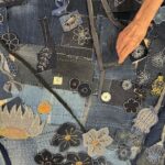 A high-angle, detailed close-up shot of a denim textile artwork, showcasing a complex and vibrant arrangement of appliquéd and embroidered floral and botanical motifs. The background is a patchwork of various shades of blue denim, with visible seams and scattered white "sashiko-style" stitching, creating a textured surface. The artwork features numerous stylized elements: - A variety of dark blue, light blue, and grey floral appliques, many outlined with white stitching and some with yellow or orange centers, are scattered across the surface. - Intricate white embroidered patterns, resembling dandelion florets and other botanical forms, are visible throughout. - Strips of denim, including sections with zippers, are integrated into the design, resembling stems or vines. - A dark blue denim patch in the upper right features a white embroidered butterfly. - In the upper right, a person's hand with slightly wrinkled skin is visible, appearing to be arranging or working on the textile. - Small white tags with numbers like "4B" are visible on some denim patches, suggesting a marking or organizational system. The image emphasizes the intricate handwork, layered construction, and the creative repurposing of denim and other materials in the textile art piece.
