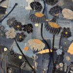 A high-angle, close-up shot captures a detailed section of a textile artwork made from various shades of blue denim. The background is a patchwork of denim scraps, with visible seams and scattered white "sashiko-style" stitching, creating a textured surface. The artwork is richly embellished with appliqué and embroidery, forming a dense field of stylized flowers and botanical motifs: - Several dark blue floral or seed pod shapes, some outlined in white, and some with orange-gold centres, are prominent. - Lighter greyish-white poppy-like flowers with textured orange or white centres also feature. - Numerous smaller flowers in different shades of blue and black are outlined with white stitching, some with yellow or orange accents. - Thin, dark blue or black fabric strips, resembling stems or vines, weave through the composition. - Delicate white embroidered leaf and floral patterns are visible throughout the darker denim sections. - Small white tags with numbers like "30" and "28" are visible near some elements, suggesting a marking or organization system. The image emphasizes the intricate handwork and the creative repurposing of denim into art.