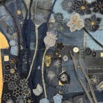 A high-angle, detailed close-up shot of a denim textile artwork, showcasing a complex and vibrant arrangement of appliquéd and embroidered floral and botanical motifs. The background is a patchwork of various shades of blue denim, with visible seams and scattered white "sashiko-style" stitching, creating a textured surface. The artwork features numerous stylized elements: - Two prominent light-coloured seed pods or poppy-like forms with textured stitching, each on a long, thin stem, are positioned vertically in the left half of the image. - A variety of dark blue and lighter blue flowers are outlined with white stitching, some with yellow or orange centres. - Intricate white embroidered patterns, resembling dandelion florets and other botanical forms, are scattered throughout. - Several strips of denim, some with zippers integrated, weave through the composition, resembling stems or vines. - A rectangular dark blue denim patch in the lower right features a detailed white embroidered butterfly. - Small white tags with numbers like "3A" and "23" are visible on some denim patches, suggesting a marking or organizational system. The image emphasizes the intricate handwork, layered construction, and the creative repurposing of denim and other materials in the textile art piece.