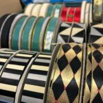 A close-up shot shows multiple spools of decorative ribbons, neatly arranged side-by-side. In the foreground on the right, there are spools of black ribbon with a harlequin pattern of alternating gold and black diamonds, accented with small red dots and a gold glitter edge. To the left, there are spools of ribbon with horizontal stripes in varying widths of black, grey, and beige. Further back and slightly out of focus, more spools are visible, including green ribbons with gold trim, a red ribbon, and ribbons with geometric patterns in black, white, and beige.