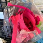 A clear plastic bag, partially open, is filled with a bright jumble of hot pink zippers and what appear to be fuchsia-colored fabric scraps or trims. The bag is placed inside a black wire mesh basket, with parts of the basket visible on the left and top.