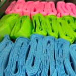 A top-down, close-up shot shows numerous rolls of brightly colored webbing or strapping neatly arranged in a green container. The rolls are organized into three distinct rows by color: the front row features vibrant light blue rolls, the middle row is a bright lime green, and the back row consists of hot pink rolls. All the rolls appear to be made of a textured, woven material.