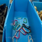 A high-angle shot captures the contents of a blue plastic bin, revealing an assortment of small, colourful carabiner clips scattered across the bottom. The carabiners come in various colours including silver, pink, black, green, and teal. To the left, another blue bin is partially visible, containing black buckles or fasteners. On the right, a third bin holds a pile of shiny, golden-coloured metal components. The background is slightly out of focus, suggesting a storage or workshop environment.