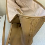 A close-up, high-angle shot focuses on the top opening and side of a light brown or tan bag, likely made of leather or faux leather. The bag has a clean, minimalist design with visible stitching along the seams and top edge. One side of the bag features a perforated pattern with small, evenly spaced holes. The interior lining of the bag appears to be a lighter, possibly cream-colored fabric, and is slightly creased. A thin strap is visible hanging down on the left side of the bag.