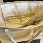 A top-down view shows the open interior of a light beige or tan tote bag, revealing its cream-colored fabric lining. On the back interior wall, there is a zippered pocket with a light gold-coloured zipper and a matching leather-like pull tab. Below the zipper, a subtle gold embroidered logo "97" is visible. The bag's outer material, also light beige, has a perforated design. Part of a light-coloured strap is seen curving across the bottom of the image. The bag is placed on a dark, possibly black, textured surface.