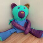 A homemade-looking stuffed bear is sitting on a light wooden floor against a cream-colored wall. The bear is made from various coloured fabrics: its head is turquoise with dark purple eyes and snout, and one ear is turquoise while the other is tan. Its torso is light blue, and its arms and legs are a mottled purple fabric with a slightly metallic sheen, though the feet are light blue with turquoise tips. A small pocket is sewn onto the bear's right side, and a piece of turquoise fabric resembling a scarf is tied around its neck.
