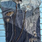 A high-angle shot captures a piece of textile artwork made from various shades of blue denim, laid flat on a light blue wooden slatted surface. The denim is patched together, with visible seams and scattered white "sashiko-style" stitching, creating a textured background. The artwork features several stylized floral and botanical elements created through appliqué and embroidery: - Two prominent dark blue, poppy-like forms with intricate white and gold stitching, resembling seed heads, are positioned in the upper half. Long, dark, textured stems extend downwards from them, with visible gold beading or wrapping on one. - Several smaller embroidered flowers with white outlines and gold or white centres are scattered across the denim. - White embroidered leaf patterns and dandelion-like motifs are also visible. The overall impression is one of detailed craftsmanship and artistic repurposing of materials, creating a stylized floral scene.