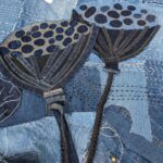 A detailed, high-angle close-up shot focuses on two large, stylized seed pods, possibly lotus or poppy heads, made from dark blue denim and positioned centrally in a textile artwork. Each seed pod features intricate appliqué and embroidery. The seed pods have dark blue bases with radiating lines of stitching. Their top surfaces are lighter blue with darker blue oval shapes, resembling seeds, appliquéd or embroidered onto them. Long, dark denim strips form the stems, extending downwards. The background is a patchwork of various shades of blue denim, from light to dark, with visible seams and abundant white "sashiko-style" dashed stitching that adds texture and a handcrafted feel. In the upper left, a small, light-coloured fabric tag is visible. The image highlights the intricate detail and layered construction of the denim art piece.