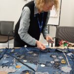 A slightly overhead shot captures a woman with curly, light-coloured hair, wearing a grey shirt and a black vest, standing over a large denim textile artwork spread on a wooden table. She is actively engaged in working on the piece, her hands holding small blue denim fabric pieces. The textile artwork is a patchwork of various shades of blue denim, intricately embellished with appliquéd and embroidered floral and botanical motifs. Several large, dark blue flowers with orange or white centres are visible in the foreground, along with lighter grey flowers. The background denim also features extensive white stitching in decorative patterns. In the background, two dark chairs are visible, and a white board or screen stands behind them. The image focuses on the artisan at work, highlighting the detailed process of creating the denim textile art.