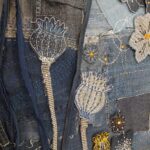 A close-up, slightly overhead shot focuses on a section of a textile artwork crafted from various shades of blue denim. The background is a patchwork of denim pieces, some with visible seams and adorned with white and light blue "sashiko-style" dashed stitching. The artwork features two prominent stylized seed pods or poppy heads. The left one is dark blue denim with intricate white stitching outlining its form and details, while the right one is a lighter blue-grey denim with similar white and blue stitching. Both have long, narrow stems made from denim. To the right, part of a larger denim appliqué flower in a light grey-blue with orange and black stitching is visible. Other smaller, darker denim flowers with yellow and orange embroidery are also scattered throughout the composition. The image highlights the detailed handwork and the textured appearance of the recycled denim.