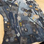 A high-angle, full shot captures a large, rectangular textile artwork laid flat on a light wooden table. The artwork is crafted from various shades of blue denim, pieced together to form a rich, textured background. The surface is extensively embellished with appliqué and embroidery, depicting a vibrant and detailed field of stylized flowers, seed pods, and botanical motifs. Key elements include: - Two large, dark blue seed pods with textured surfaces in the upper left, and lighter greyish-white poppy-like flowers with orange or black centres in the upper right. - Numerous smaller flowers in different shades of blue and black, outlined with white stitching, and accented with orange or yellow, are scattered throughout the composition. - Thin, dark stems made from denim strips or embroidery connect many of the floral elements. - The lower sections feature a dense array of intricate white embroidered patterns, resembling small flowers, leaves, and abstract botanical forms, including dandelion-like motifs. - Several small white tags with numbers like "3A", "23", and "4B" are visible on some denim patches, suggesting a marking or organizational system. - A rectangular dark blue denim patch in the lower right features a white embroidered butterfly. The overall impression is one of detailed craftsmanship and artistic repurposing of materials, showcasing the entire elaborate denim art piece before it is mounted.