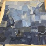 A high-angle shot captures a large, rectangular piece of fabric laid flat on a light-coloured table, entirely covered with a patchwork of various shades of blue denim scraps. The denim pieces are of different sizes and textures, some with visible seams, pockets, or faded areas. In the lower centre of the patchwork, a pair of silver-handled scissors is resting on a dark denim patch. Nearby, a dark denim patch with a white embroidered floral outline is visible. Other pieces of denim also feature subtle embroidery or patterns, hinting at their original use. The background shows a dark blue carpet. Part of a chair and a white surface are visible at the top. The image depicts the initial stage of a denim textile project, showcasing the raw materials before assembly.