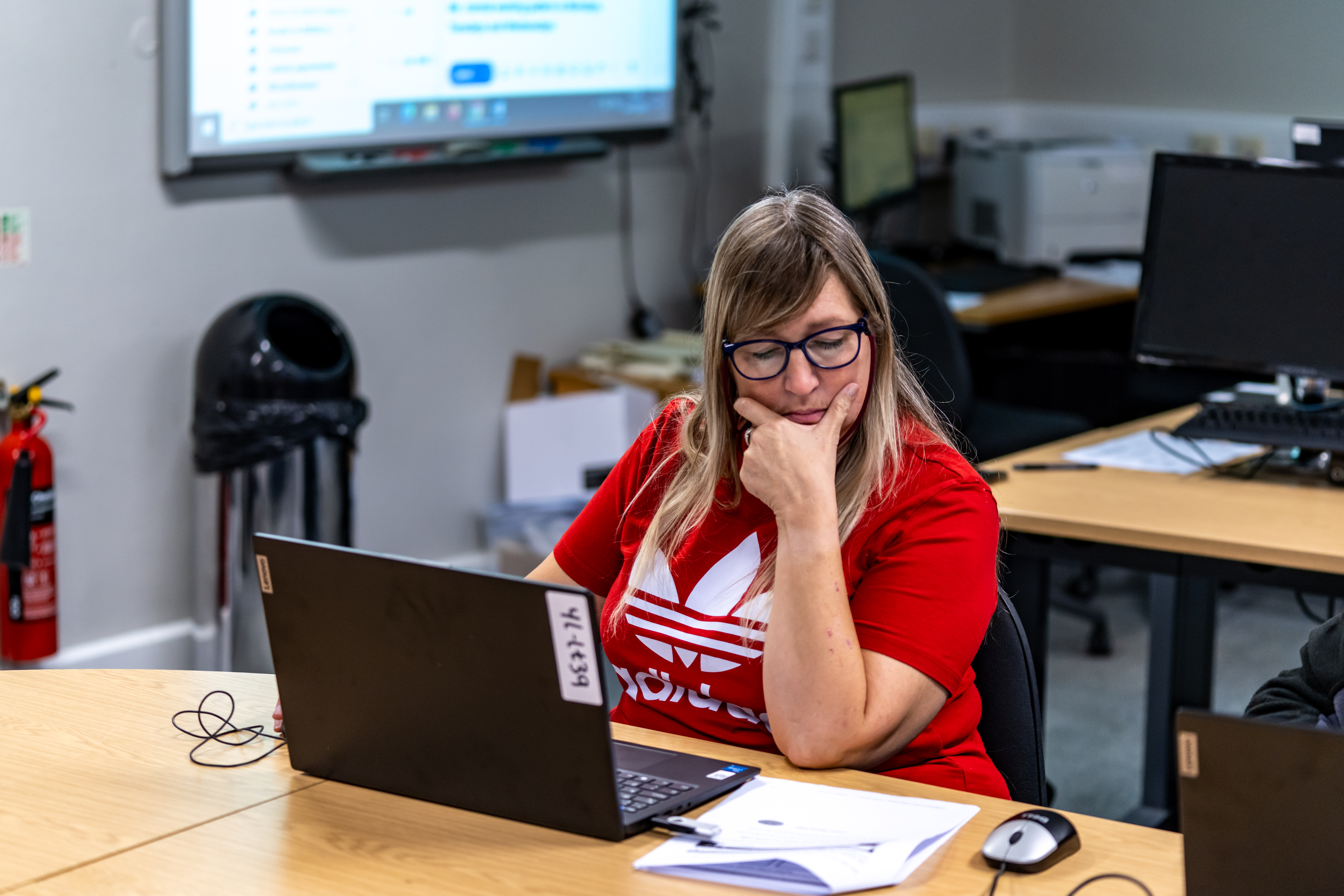 A learner in a digital skills class, sitting at a laptop and reading a page of guidance notes next to her on the desk.
