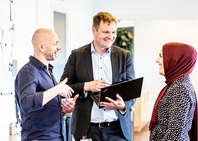 Three colleagues chatting and smiling in an office, one holding an open laptop, with a whiteboard visible in the background.